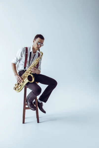 Hermoso joven jazzman sentado en taburete y jugando saxofón en gris - foto de stock
