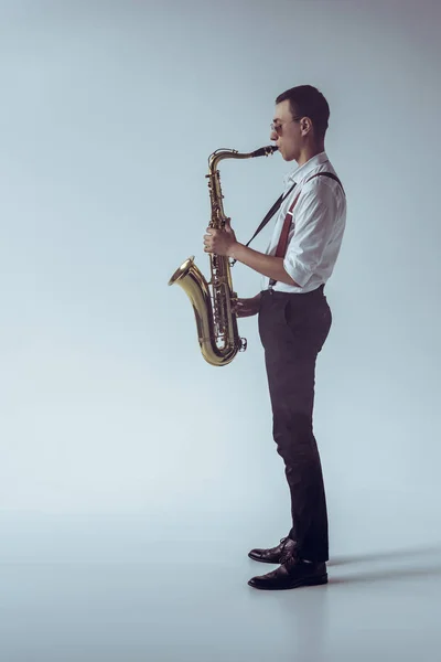 Vista lateral de un joven músico profesional con estilo tocando el saxofón en gris — Stock Photo