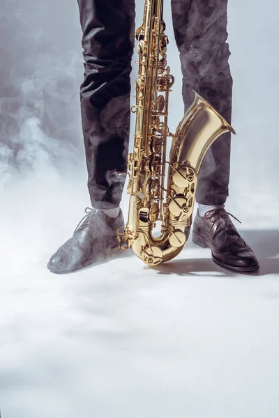 Частичный взгляд молодого музыканта, стоящего с саксофоном в дыму на сером — стоковое фото