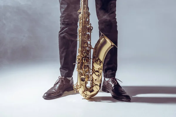 Vista parcial del músico profesional parado con saxofón en humo sobre gris — Stock Photo
