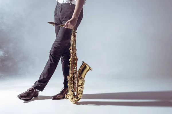 Обрезанный снимок профессионального музыканта, стоящего с саксофоном в дыму на сером — стоковое фото
