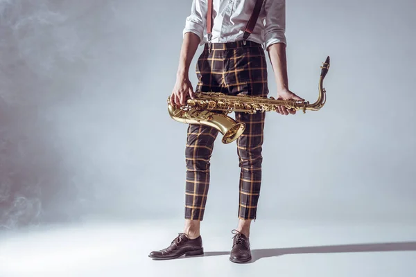 Cropped shot of stylish young musician standing and holding saxophone in smoke on grey — Stock Photo