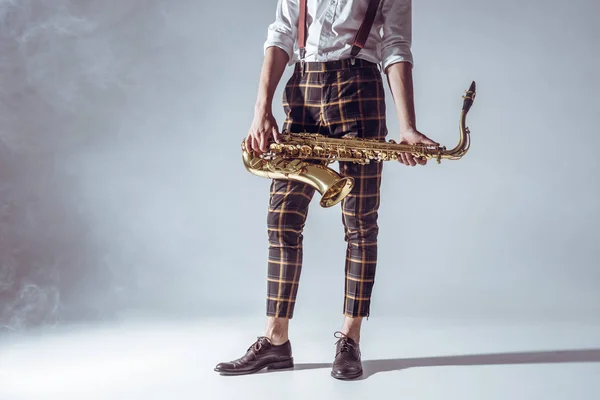 Recortado disparo de elegante joven músico de pie y la celebración de saxofón en el humo en gris - foto de stock