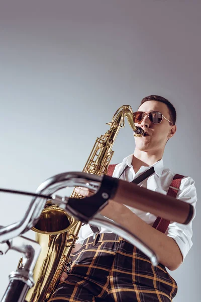Vue de bas angle de jeune performer élégant dans les lunettes de soleil jouant saxophone sur gris — Photo de stock