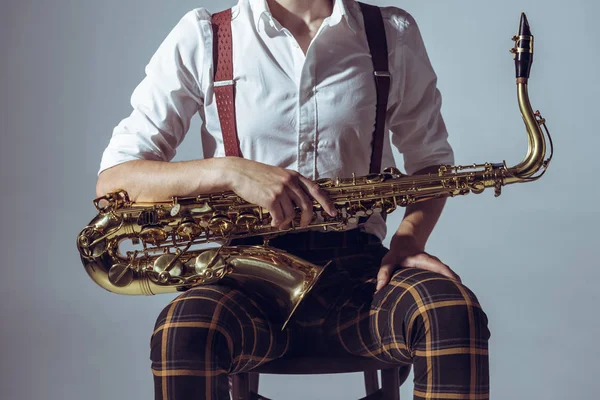 Обрезанный снимок молодого музыканта, сидящего на стуле и держащего саксофон, изолированный на сером — стоковое фото