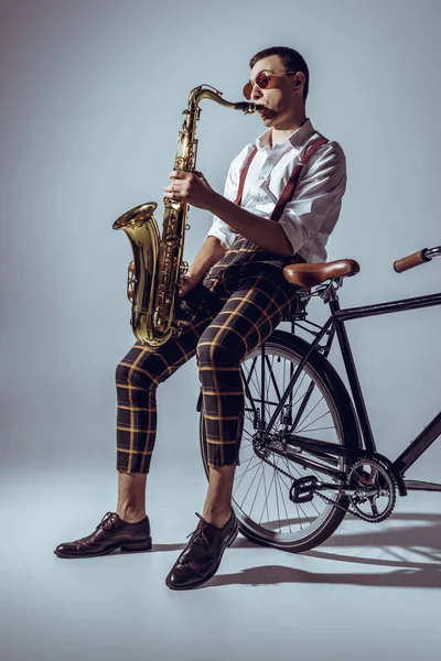 Guapo joven intérprete en gafas de sol jugando saxofón mientras está sentado en bicicleta en gris — Stock Photo