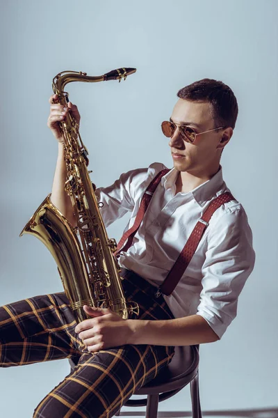 Beau jeune performer dans des lunettes de soleil tenant saxophone et regardant loin sur gris — Photo de stock