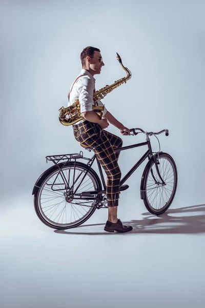 Young stylish musician in sunglasses holding saxophone and riding bicycle on grey — Stock Photo