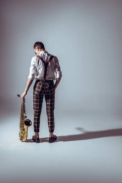 Vista posteriore di elegante giovane musicista in piedi con sassofono su grigio — Foto stock