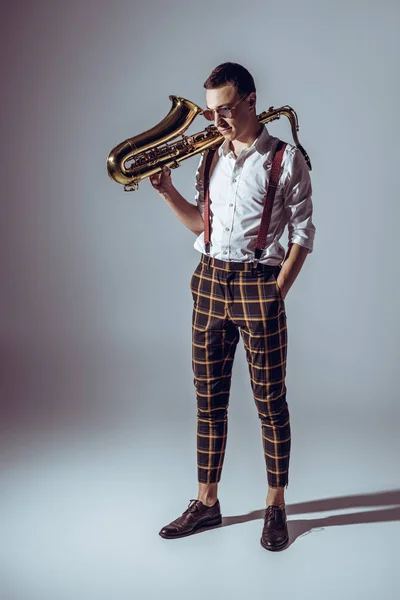 Стильный молодой музыкант в солнечных очках держит саксофон и смотрит свысока на серый — стоковое фото