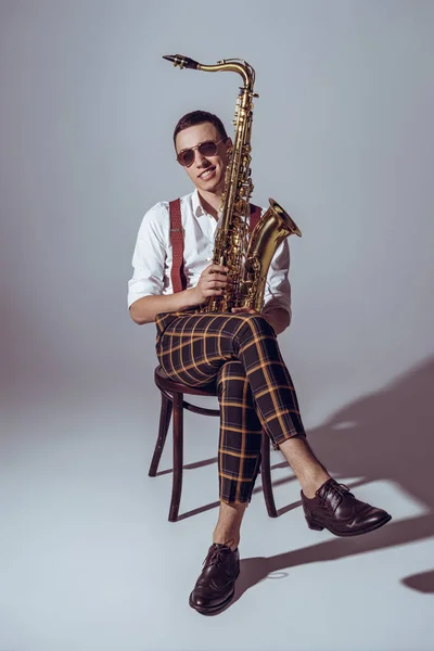 Apuesto joven saxofonista en gafas de sol sonriendo a la cámara mientras está sentado con saxofón en gris - foto de stock