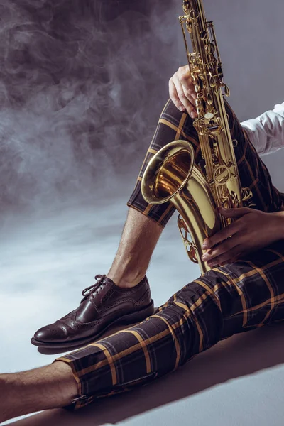 Vista parcial del joven músico elegante sentado con saxofón en humo sobre gris - foto de stock