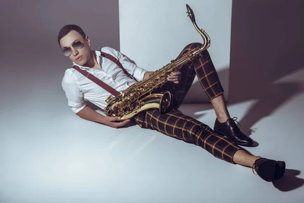 Beau jeune musicien élégant en lunettes de soleil couché avec saxophone sur gris — Photo de stock