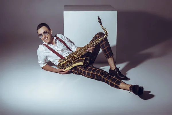 Smiling young musician in sunglasses holding saxophone while lying on grey — Stock Photo