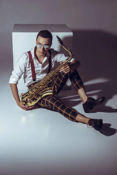 Stylish young musician in sunglasses sitting with saxophone on grey — Stock Photo