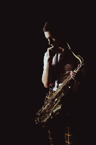 Pensativo joven músico sosteniendo saxofón aislado en negro - foto de stock