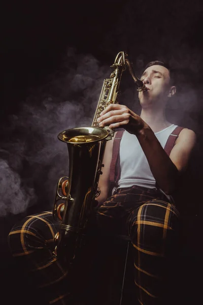 Vista de bajo ángulo de expresivo joven músico tocando el saxofón en el humo en negro - foto de stock