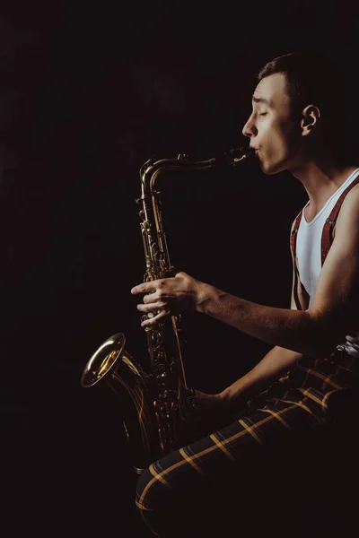 Vista lateral de saxofonista joven elegante sentado y jugando saxo en negro - foto de stock