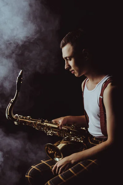 Vue latérale du jeune jazzman élégant assis et tenant le saxophone en fumée sur noir — Photo de stock