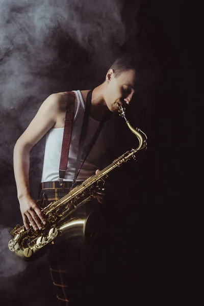 Handsome young musician playing saxophone in smoke on black — Stock Photo