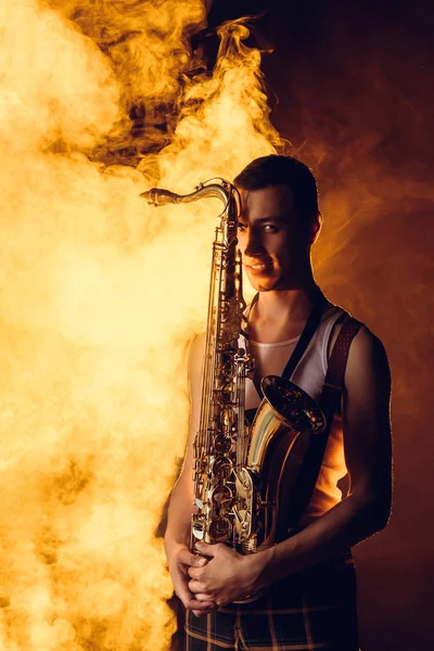 Apuesto joven músico elegante sosteniendo saxofón y mirando a la cámara en el humo - foto de stock