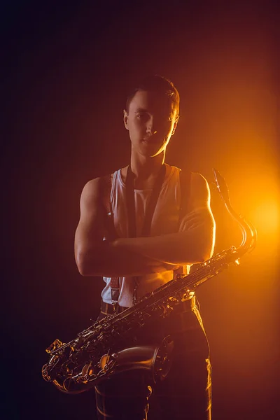 Joven músico guapo con saxofón de pie con los brazos cruzados y mirando a la cámara — Stock Photo