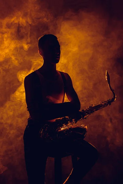 Silhouette of young musician sitting on stool with saxophone in smoke — Stock Photo