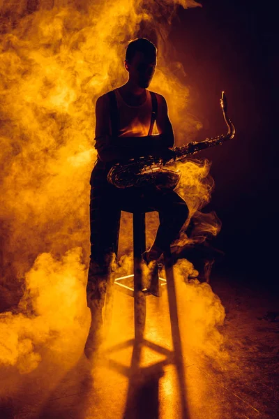 Silueta del joven músico sentado en el taburete y sosteniendo el saxofón en el humo - foto de stock