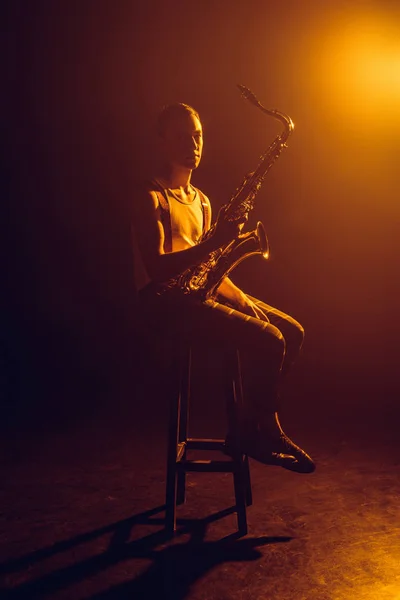Молодий джазмен дивиться на камеру, сидячи на табуреті і тримаючи саксофон — стокове фото