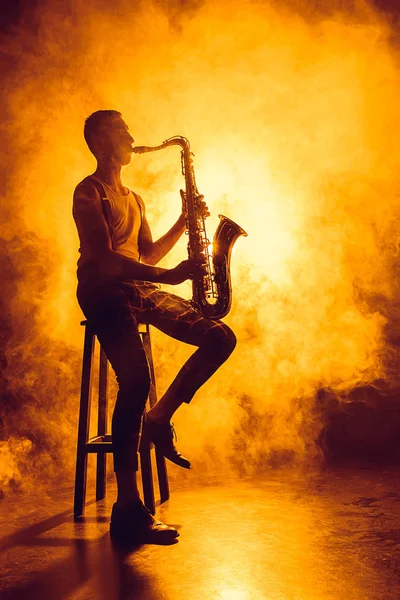 Joven músico profesional sentado en el taburete y tocando el saxofón en el humo - foto de stock