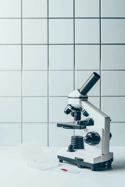 Muestra de sangre y microscopio óptico en comprimidos blancos frente a la pared de azulejos - foto de stock