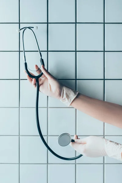 Inyección recortada de médico en guantes que sostienen estetoscopio frente a la pared blanca de azulejos - foto de stock