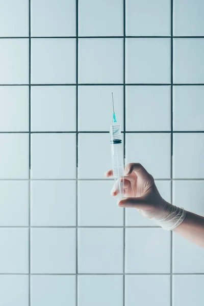 Cropped shot of doctor in glove holding syringe with transparent liquid in front of tiled white wall — Stock Photo