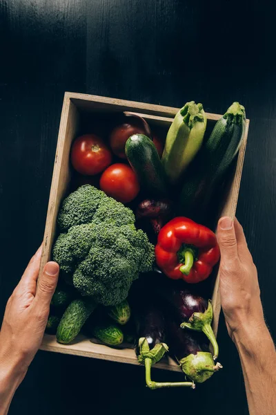 Обрізане зображення людини, що тримає дерев'яну коробку з овочами над темним столом — стокове фото