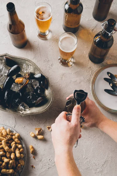 Cropped shot of man opening bottle of cold beer at tabletop with peanuts and mussels — Stock Photo