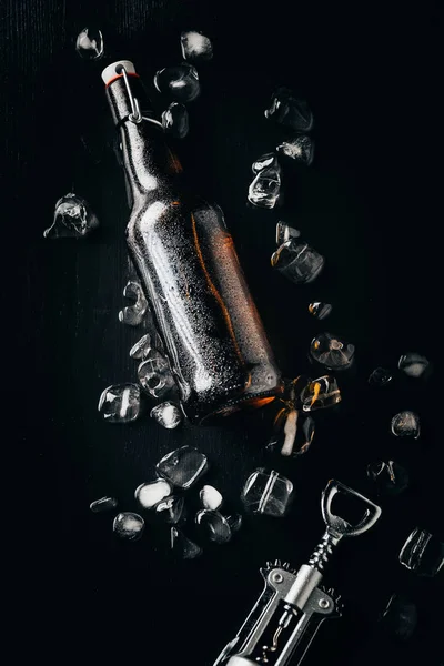 Flat lay with bottle opener, bottle of beer on ice cubes arranged on dark tabletop — Stock Photo