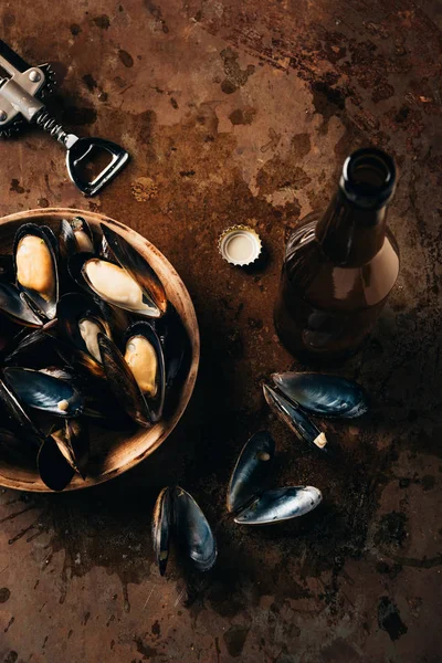 Flat lay with opened glass bottle of beer, bottle opener and mussels in bowl on rust surface — Stock Photo