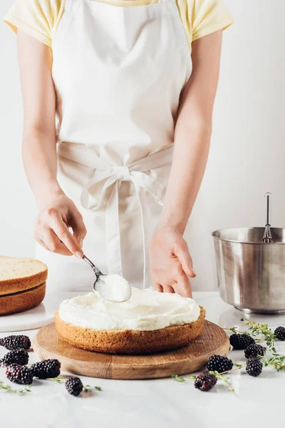 Tiro recortado de mujer en delantal blanco aplicando crema sobre pastel recién horneado en blanco — Stock Photo