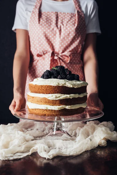 Cropped shot of woman holding freshly baked blackberry cake on glass stand on black — Stock Photo