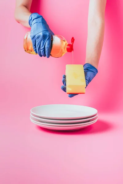 Imagen recortada de la mujer en guante de goma verter líquido lavavajillas en la esponja de lavado sobre la pila de platos blancos, fondo rosa - foto de stock