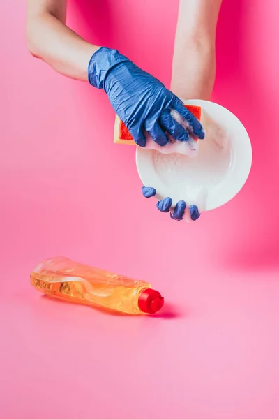 Imagem cortada de limpador feminino em placa de lavagem de luvas de borracha perto de líquido de lavagem de louça, fundo rosa — Fotografia de Stock