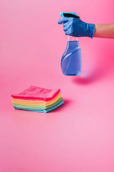 Imagem cortada de limpador feminino segurando fluido de limpeza perto da pilha de trapos coloridos, fundo rosa — Fotografia de Stock