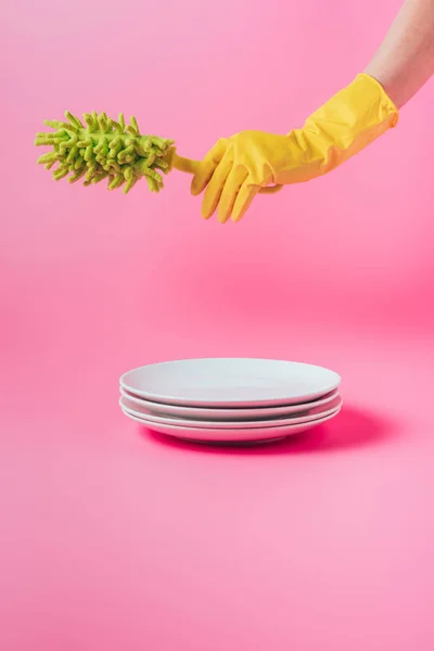 Cropped image of woman in rubber glove holding dish microfiber brush over stack of white plates, pink background — Stock Photo