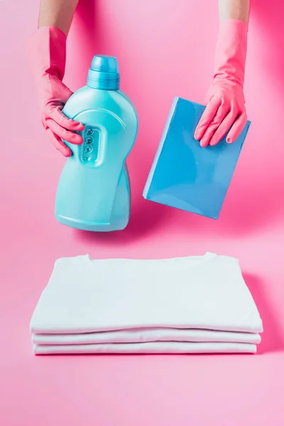 Vista parcial del limpiador femenino en guantes de goma que sujetan el líquido de la ropa y el polvo de lavado sobre una pila de camisetas blancas limpias, fondo rosa - foto de stock