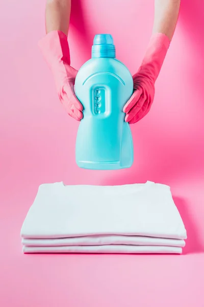 Imagem cortada do limpador fêmea em luvas de borracha segurando líquido de lavanderia sobre pilha de camisetas brancas limpas, fundo rosa — Fotografia de Stock