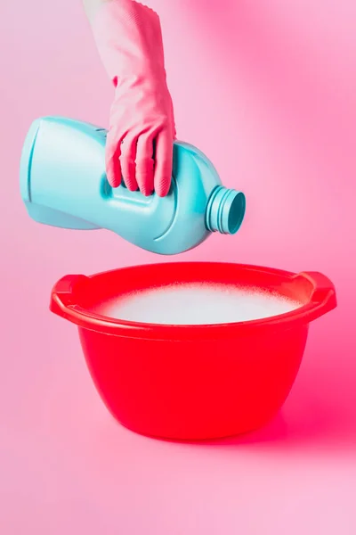 Vista parcial del limpiador femenino vertiendo líquido de lavandería en lavabo de plástico con espuma, fondo rosa - foto de stock