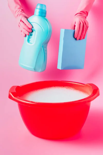 Cropped image of female cleaner in rubber gloves holding laundry liquid and washing powder over plastic basin with foam, pink background — Stock Photo