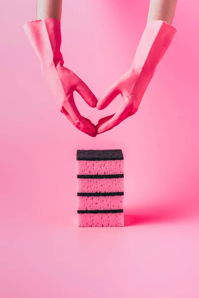 Imagen recortada de la mujer en guantes de goma haciendo símbolo del corazón por las manos sobre la pila de esponjas de lavado, fondo rosa - foto de stock