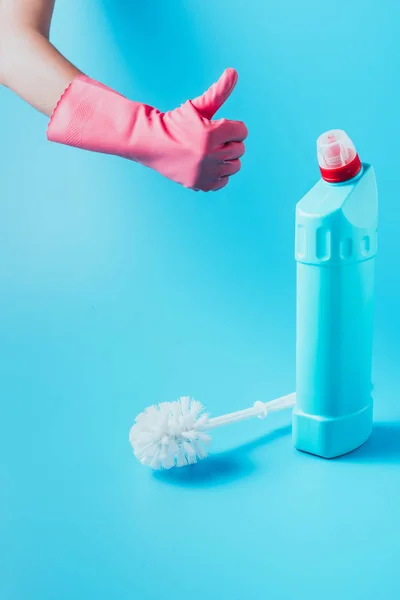 Imagen recortada de limpiador femenino en guante de goma haciendo gesto de golpe hacia arriba cerca de líquido de limpieza con cepillo de inodoro, fondo azul - foto de stock