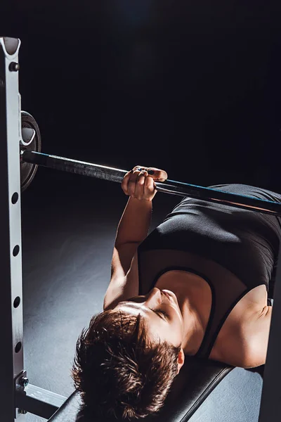Vista de ángulo alto de la deportista haciendo ejercicio con barra de pesas en el gimnasio, fondo negro - foto de stock